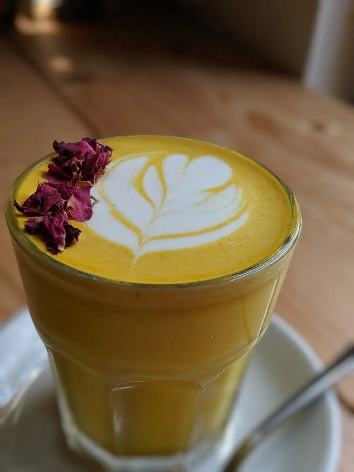 A turmeric-yellow latte in a glass with rosette latte art and dried rose petals, on a saucer on a wood table.