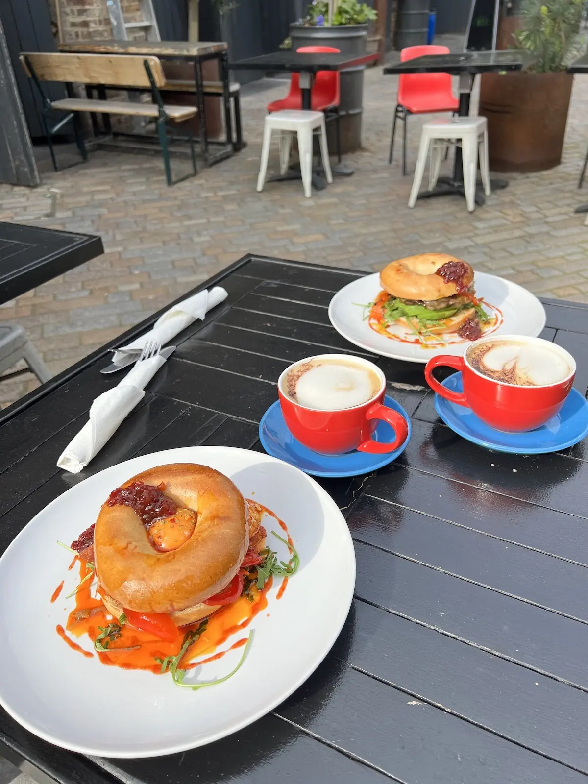 An outdoor pavement table set with two bagels and two red cups of cappuccino, terrace seating behind.