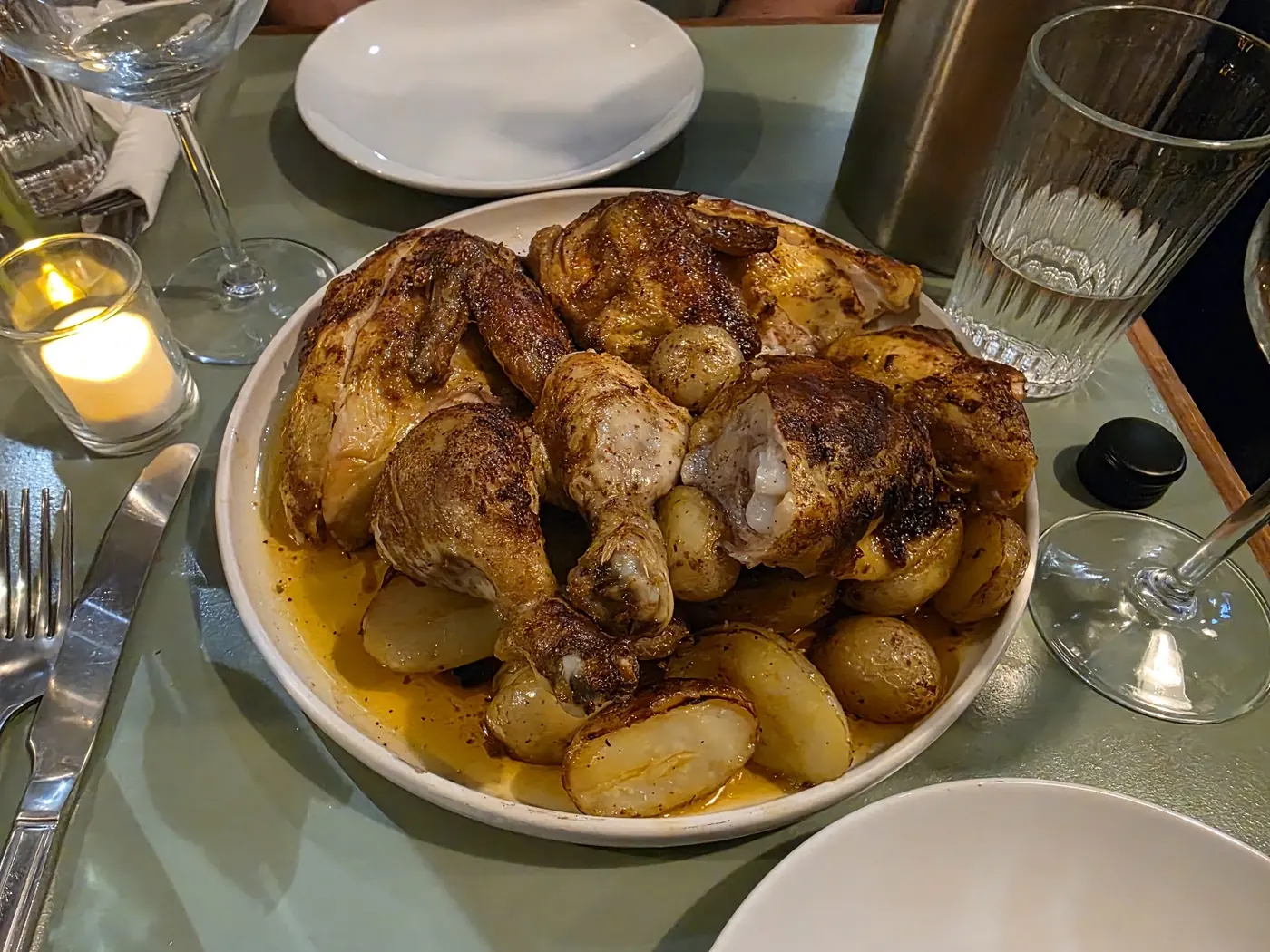 A bowl of golden roast chicken pieces with roast potatoes, on a green table lit by a candle.